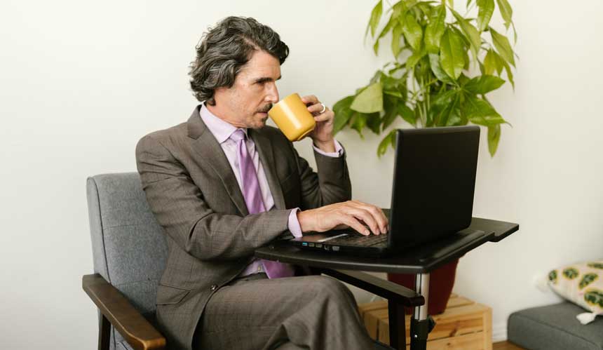 man sitting on chair holding yellow mug to drink coffee while working on his laptop